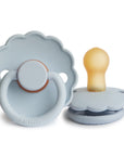 The FRIGG Daisy Natural Rubber Pacifier 2-Pack in Powder Blue features two scalloped-edge pacifiers—one upright displaying the circular handle, the other on its side—with yellow natural rubber latex nipples.