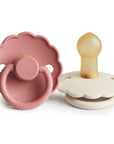 The FRIGG Daisy Natural Rubber Pacifier 2-Pack includes two scalloped pacifiers: one pink with a round handle (front view) and one cream featuring a natural rubber latex nipple (side view).