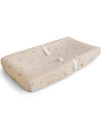 The mushie Extra Soft Muslin Changing Pad Cover is shown on a beige, contoured pad with small brown dots and two white safety straps, all displayed against a white background.