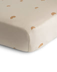 A close-up of the mushie Stretchy MINI Crib Sheet in light beige, adorned with small brown minimalist rainbows. Made from breathable organic cotton, the sheet fits snugly around a mini crib mattress corner.