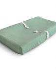 The mushie Extra Soft Muslin Changing Pad Cover in light green is contoured for comfort and includes two white safety straps, displayed on a white background.