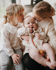 Three children sit together as an older girl cradles a baby holding the mushie No-Drop Sensory Teether. Another girl sits nearby, gently kissing the baby's head, their light clothing and closeness creating a peaceful, heartwarming moment.