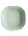 A light green, square-shaped Square Dinnerware Bowl by mushie, featuring rounded edges and the brand’s name embossed in the center, made from BPA-free plastic, is photographed on a white background.
