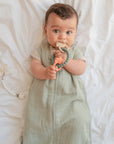 A baby in a mushie Organic Cotton Sleep Bag rests on a white bed, holding and chewing a teething ring, with a pacifier attached to a beaded clip lying nearby.
