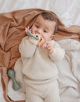 A baby in a cream knit outfit lies on beige and tan blankets, holding and chewing on the mushie Dino Teething Ring. A green sensory toy rattle is nearby.