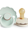 The FRIGG Daisy Natural Rubber Pacifier 2-Pack by FRIGG features two pacifiers with scalloped edges—one in soft mint green (upright) and one in off-white (sideways)—both with natural rubber latex nipples.
