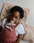 A young child with curly hair lies on a pink and beige checkered mat, wearing a white shirt and pink overalls, looking up with a pacifier held securely by the mushie Linen Pacifier Clip 2-Pack.