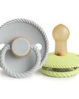 The Mushie FRIGG Rope Natural Rubber Pacifier 2-Pack includes one light gray pacifier with a ring handle and one light green, both featuring a rope-textured edge and latex nipple. Both stand upright on a white background.
