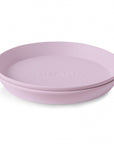 A light purple, round mushie Round Dinnerware Plate with raised edges and "mushie" embossed in the center, made from non-toxic plastic, is shown on a white background.