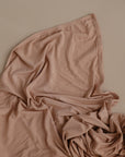The mushie Stretchy Swaddle, in soft light brown, is gently spread out on a matching beige surface, forming subtle folds and textured lines.