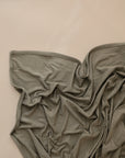 The mushie Stretchy Swaddle, a soft olive-green breathable baby blanket, is loosely spread and wrinkled on a smooth, light beige surface.