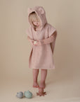 A small child wearing a soft pink hooded towel with bear ears stands barefoot on a beige background, looking down and holding a toy, with three mushie Sealife Mold Free Bath Play Set silicone toys on the floor in front of them.