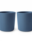 Two simple, cylindrical mushie Dinnerware Cups in blue are placed side by side on a white background.