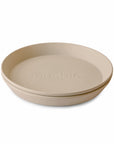 A beige, round mushie Round Dinnerware Plate with raised edges and 'mushie' embossed in the center is shown against a white background. Made from non-toxic, dishwasher-safe plastic.