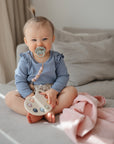 A baby with a pacifier sits on a beige sofa, wearing a blue long-sleeve top and holding a pastel wooden toy palette. Nearby are a pink blanket and mushie Organic Cotton Muslin Cloths 3-Pack, with neutral cushions in the background.