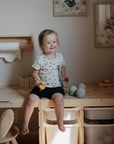 A young child with blonde hair sits on a wooden table in a cozy room, holding a toy and the mushie Linen Pacifier Clip 2-Pack. The background shows framed animal art, drawing paper, and organized storage bins.