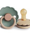 The FRIGG Daisy Natural Rubber Pacifier 2-Pack features two daisy-shaped pacifiers with scalloped edges—one upright in muted green and beige, one lying down showing the natural rubber nipple. Both have round handles embossed with "FRIGG.