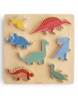 The Mushie Wooden Puzzle includes seven colorful dinosaur pieces—pterodactyl, triceratops, stegosaurus, spinosaurus, brachiosaurus, tyrannosaurus, and plesiosaur—that help kids build fine motor skills as they fit them onto a light wood board.