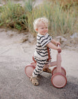 Toddler outdoors in striped outfit with mushie Linen Pacifier Clip 2-Pack, smiling on pink bike.