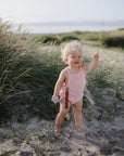 Smiling toddler with mushie Linen Pacifier Clip 2-Pack stands on sandy path, pointing up.