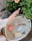 A young child in striped clothing plays outdoors, holding mushie's Space Mold Free Bath Play Set above a white basin filled with water and other mold-free bath toys, surrounded by green plants and blooming flowers.