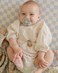 A baby in a light romper smiles on a checkered blanket, holding its feet. A mushie Organic Cotton Muslin Swaddle Blanket with floral pattern and a toy surround them; their pacifier is clipped to the outfit.