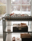 A baby rests on the mushie Extra Soft Muslin Changing Pad Cover atop a changing table by sunlit windows, with baskets of supplies below. Outside, snow blankets the ground and sunlight softly warms the cozy room.