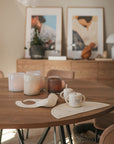 A wooden table with the Bluey Gift Set by mushie, candles, mugs, and art in the background.