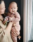 A woman holds a baby dressed in mushie's pink Two Piece Set by a window. The baby faces the camera, holding a toy ring and pacifier clip, bathed in soft natural light through the curtains.