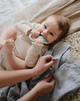 Baby chews Mushie Animal Ring Teether as an adult fastens their blue garment on a blanket.