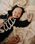 A smiling baby wearing a black skeleton onesie lies on a bed, holding the mushie Key Toy teether to soothe their gums, while an adult hand nearby holds a wooden spatula.