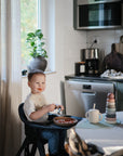 A young child with food on their face smiles in a high chair, eating from the Mushie Classic Silicone Suction Plate. A cup and colorful stacking toy sit nearby on the table as sunlight streams through a window with a potted plant.