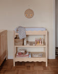 A wooden changing table with folded towels, baby care items, toys, a mushie Sun Teether, and a checkered cloth stands next to a crib in a cozy nursery with wooden flooring and a smiling face wall decoration above.