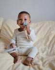 A smiling baby in cream pajamas sits on a bed with light yellow bedding, contentedly holding the FRIGG Andersen Fairytale Silicone Pacifier 2-Pack by FRIGG, made in Denmark, and a teething toy, looking playful.