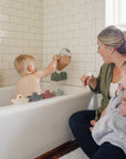 A woman sits by the tub holding a baby in a towel, while a toddler plays and pours water. mushie Bath Boats, made from non-toxic plastic, are stacked along the tub's edge.