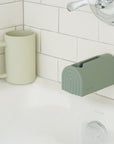 A light green cup rests on a white tile bathtub edge beside the Mushie Spout Cover, a gray-green silicone cover with an arched design, as water pours into a bubble-filled tub for safe and fun bath time.