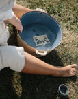 A child sits barefoot on grass next to a large blue pot of water, holding the mushie Sun Teether. A round container lid rests on the ground nearby.