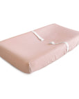 The mushie Extra Soft Muslin Changing Pad Cover fits a light pink contoured pad with raised sides and two white safety straps, offering ultimate comfort for your baby.