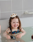 A young child in a blue swimsuit smiles in the tub with wet hair, balancing a mushie Bath Boat on their head as other water toys float nearby. White tiles and a grab bar are visible.