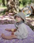 A baby with a Mushie FRIGG Rope Natural Rubber Pacifier sits on a purple daisy-print blanket outdoors, holding a wooden toy. Pink petals are scattered on the ground, and a bicycle is blurred in the background—a sweet baby moment.