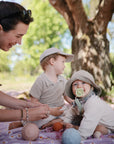 A woman sits outdoors on a blanket with two young children, surrounded by soft toys and baby essentials. The baby, wearing a sunhat, uses the Mushie FRIGG Rope Natural Rubber Pacifier 2-Pack and looks at the camera. The other child in a cap looks away.