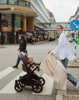 Smiling woman pushes stroller; baby wrapped in mushie Organic Cotton Muslin Swaddle Blanket.