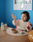 A young child wearing a bib sits at a wooden table, smiling with raised hands. In front of them are a bowl, food plate, bottle, and the mushie Silicone Placemat. The background has a blue wall and a framed picture.