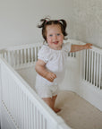 Toddler smiles in white crib on mushie Extra Soft Muslin Crib Sheet, holding the rail.