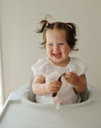 Smiling toddler in high chair holds food and a mushie Linen Pacifier Clip 2-Pack.