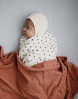 Baby swaddled in mushie Organic Cotton Muslin Swaddle Blanket on rust cloth on a gray surface.