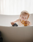 A young child smiles in a white bathtub, playing with mushie’s Vehicles Mold Free Bath Play Set—gray and yellow silicone toys—near the tub’s edge as soft light streams in through a window behind them.