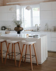Modern white kitchen with an island and three wooden stools, minimal decor, a large vase with greenery, pendant lights above, and a mushie Lunchbox with built-in compartments placed by the window.
