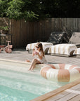 Girl by pool, feet in water, wearing sunglasses, with a mushie Mini Toddler Bike beside her.