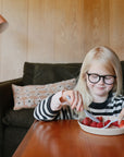 A young girl with blonde hair and glasses smiles as she cuts strawberries at a wooden table using the mushie Dinnerware Cutlery Set, a non-toxic, dishwasher-safe set. She wears a black-and-white striped sweater; a sofa and lamp are visible behind her.
